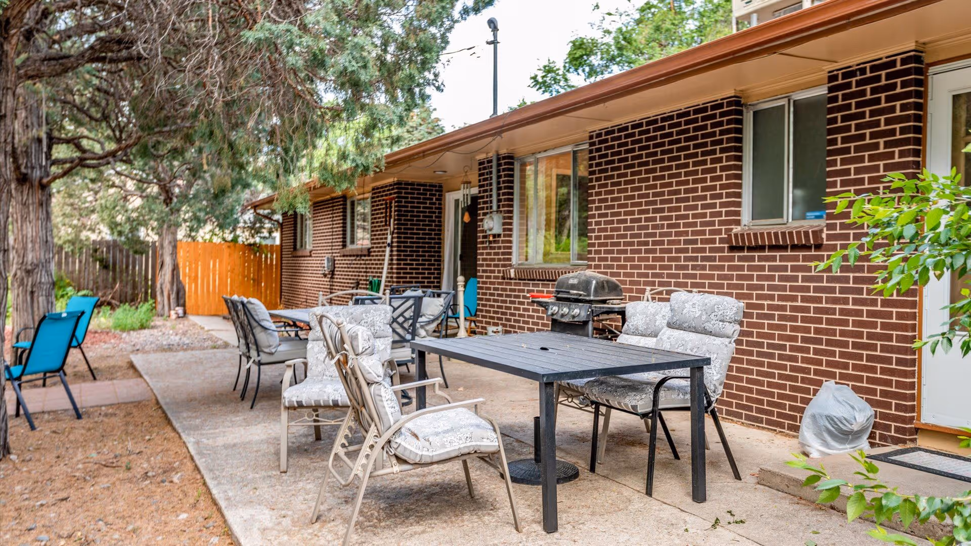 Outdoor patio area with several cushioned chairs and tables set on a concrete surface next to a brick building. There is a barbecue grill near the building wall, trees providing shade, and a wooden fence in the background.