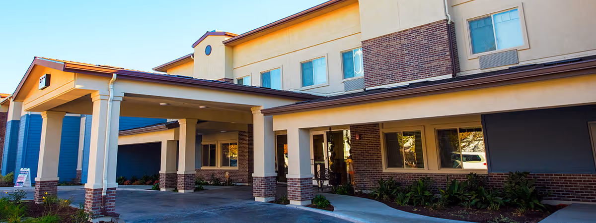 Exterior view of Summerset Senior Living facility showing the entrance with a covered drop-off area supported by columns. The building features a combination of beige stucco and red brick with multiple windows and a clear blue sky above.