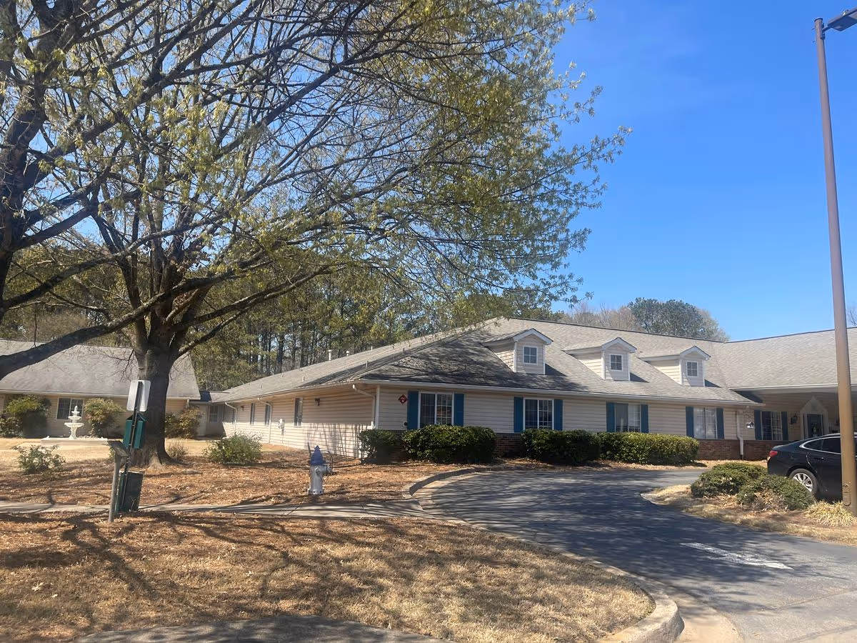 Front exterior of a single-story senior living building with a curved driveway, lawn and large tree under a clear blue sky.