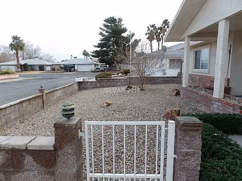 Front yard of a single-story house with a gravel-covered garden area enclosed by a low brick wall and a white metal gate. There are some small bushes near the house and a few leafless trees in the yard. The street and neighboring houses are visible in the background under an overcast sky.