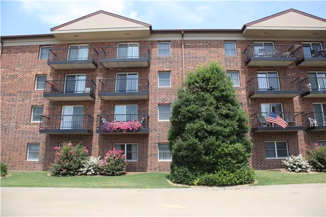 Exterior view of a three-story brick apartment building with balconies, some decorated with flowers and an American flag. There is a large green tree and bushes in front of the building under a partly cloudy sky.