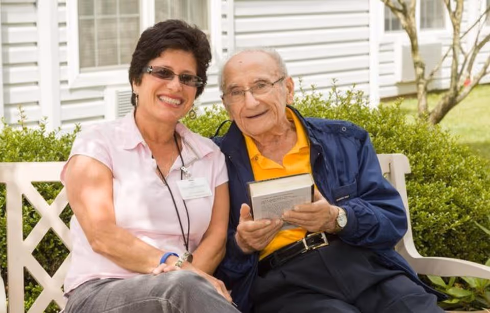An elderly man holding a book sits on a white bench outside next to a smiling woman wearing glasses and a name badge. They are in a garden area with green bushes and a white building with windows in the background.