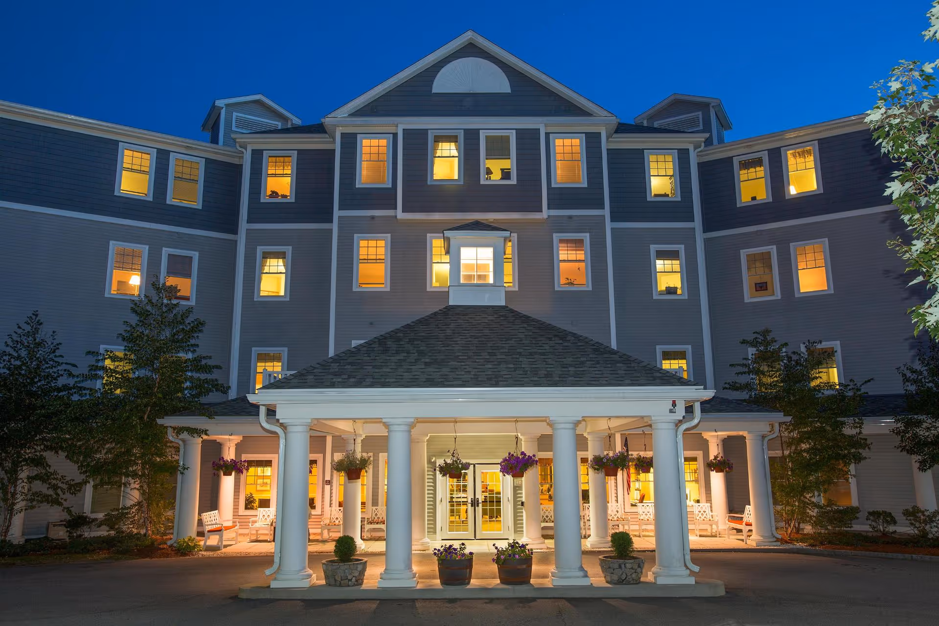 A large multi-story senior living building at dusk with illuminated windows and a columned entrance.