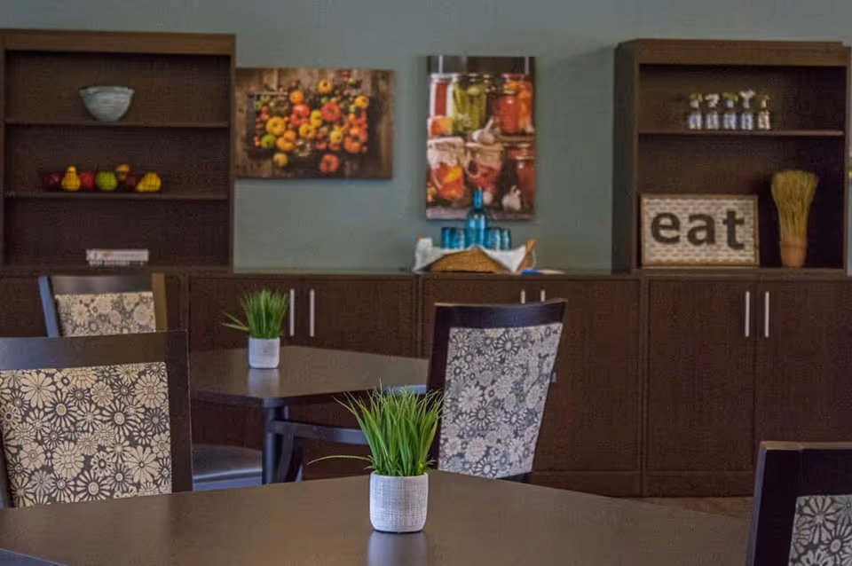 Interior view of a dining area with dark wooden tables and chairs featuring floral patterned upholstery. Small potted plants are placed on the tables. In the background, there are dark wooden cabinets and shelves with decorative items including a bowl, artificial fruits, jars, and a sign that says 'eat'. Two colorful food-themed paintings hang on the wall above the cabinets.