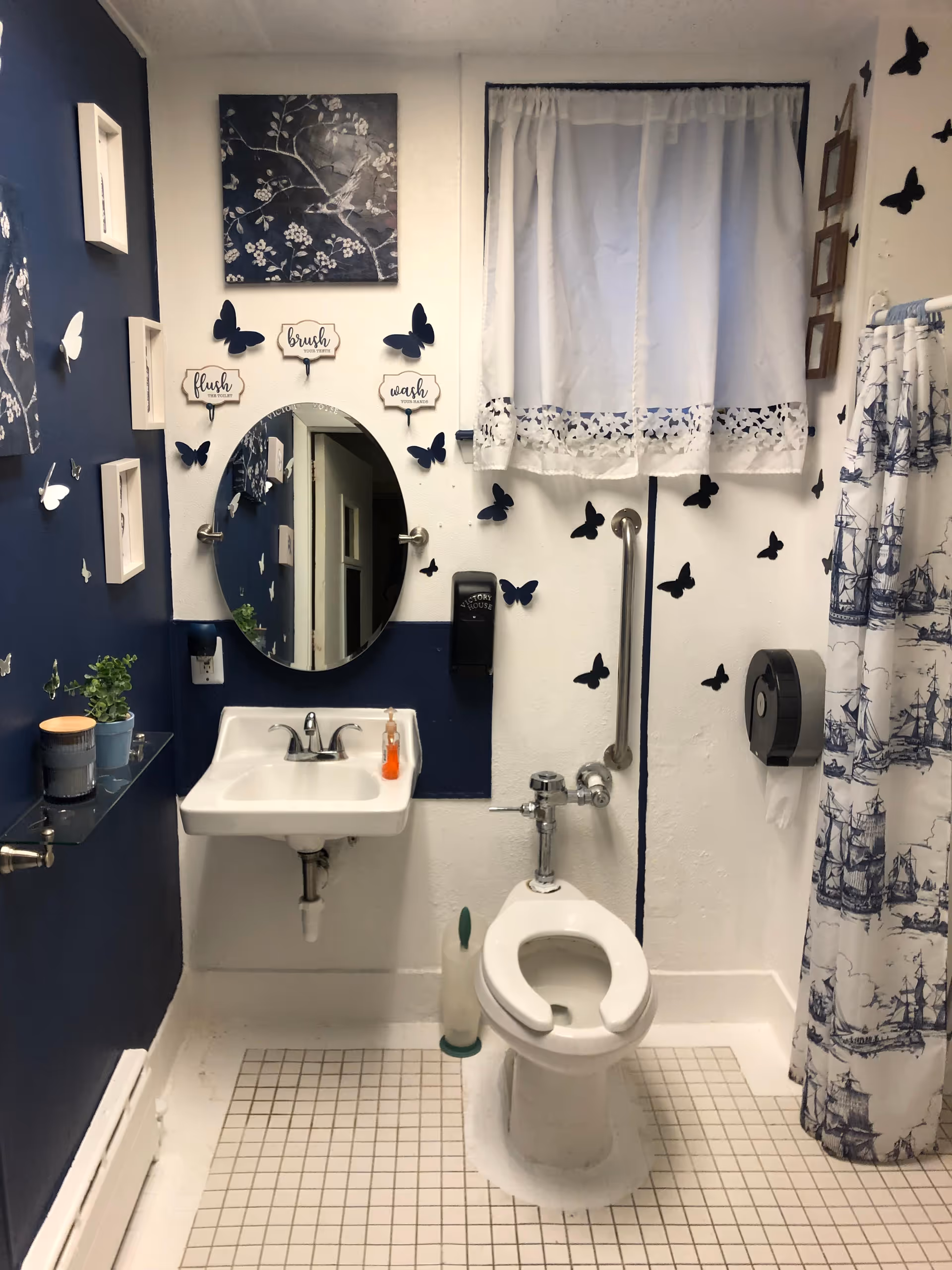 Small bathroom with blue walls, butterfly decor, round mirror, sink, toilet, shower curtain, grab bar.