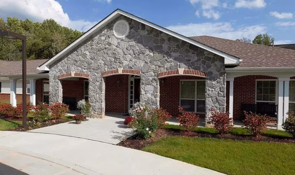 Exterior view of a senior living facility building with stone and brick facade, arched entryways, landscaped garden beds with shrubs and flowers, and a clear blue sky with some clouds.