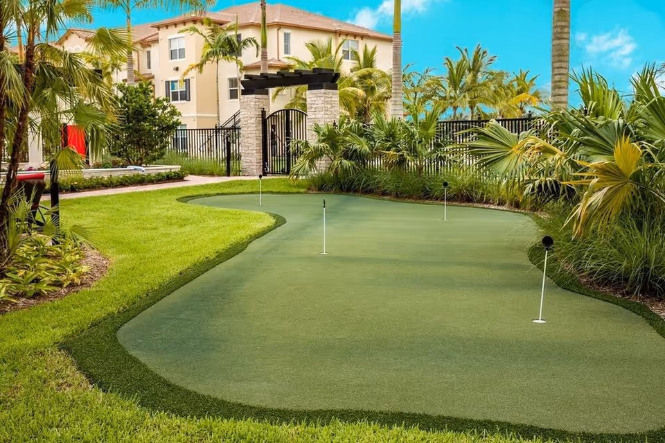 A landscaped outdoor putting green surrounded by palm trees and a building in the background.
