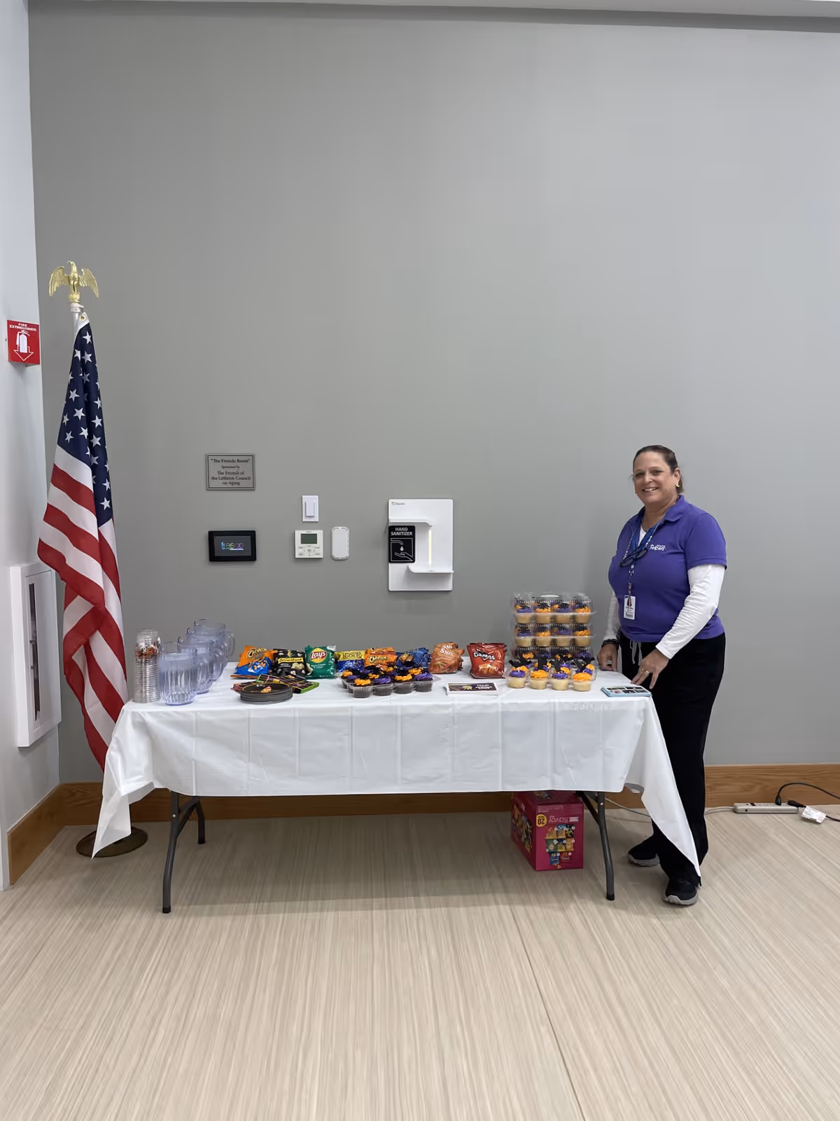 Staff member in purple shirt stands beside a snack table indoors.