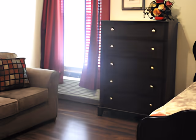 Small furnished room with a couch, a tall dark dresser, the edge of a bed, and a window with red curtains.