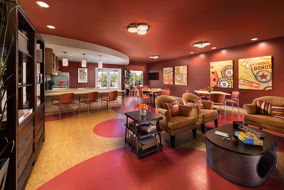 A spacious senior living lounge with red walls, comfortable seating, and a service bar.
