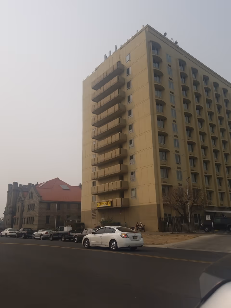 Tall beige multi-story building with stacked balconies and a 'SUN TOWER' sign, cars parked along the street in front.
