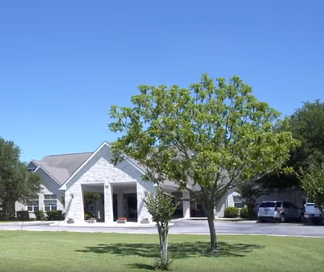 Front exterior of Argent Court Assisted Living showing the covered entrance, lawn, and a tree under a clear blue sky.