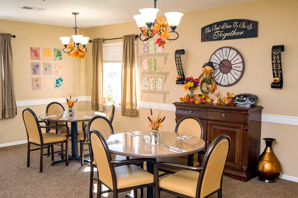 A cozy dining room with two round tables, each surrounded by four chairs with beige cushions. The tables are set with silverware and small floral centerpieces. The room has beige walls with decorative artwork, a large clock, and a sign that reads 'The Best Place To Be Is Together.' There is a wooden sideboard decorated with autumn-themed items and a small stereo. Two windows with beige curtains allow natural light to brighten the space.