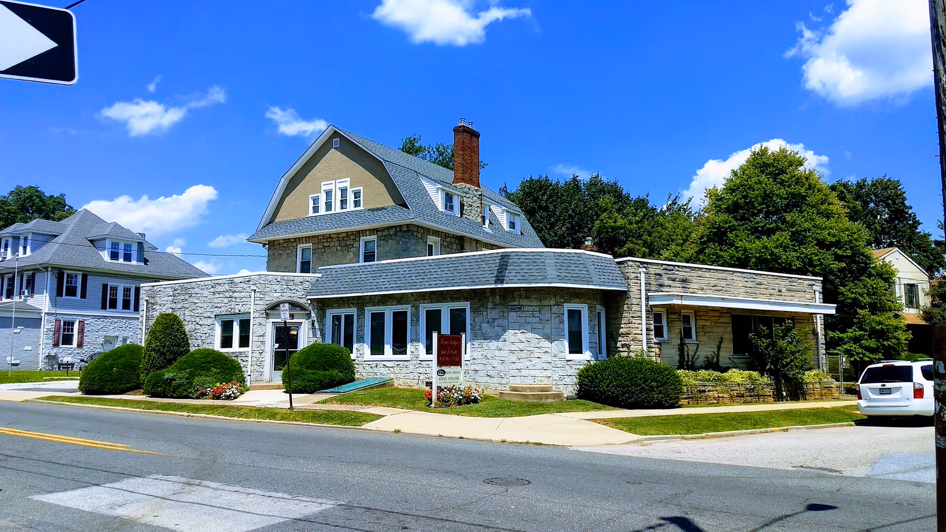 Stone building on a quiet street with manicured shrubs, clear blue sky, and a parked car.