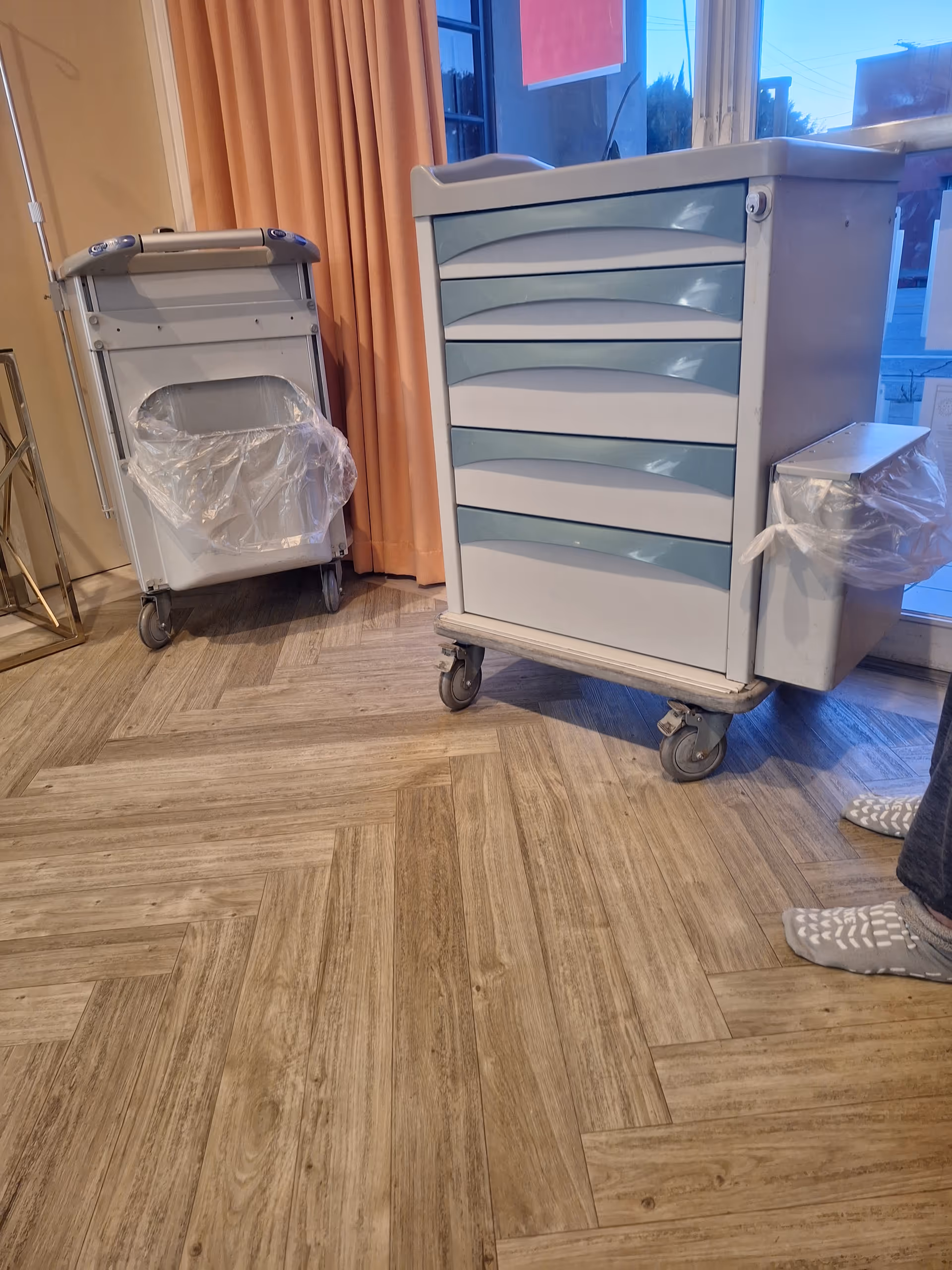 Medical carts on wheels and a plastic-wrapped bin near orange curtains in a clinical room.