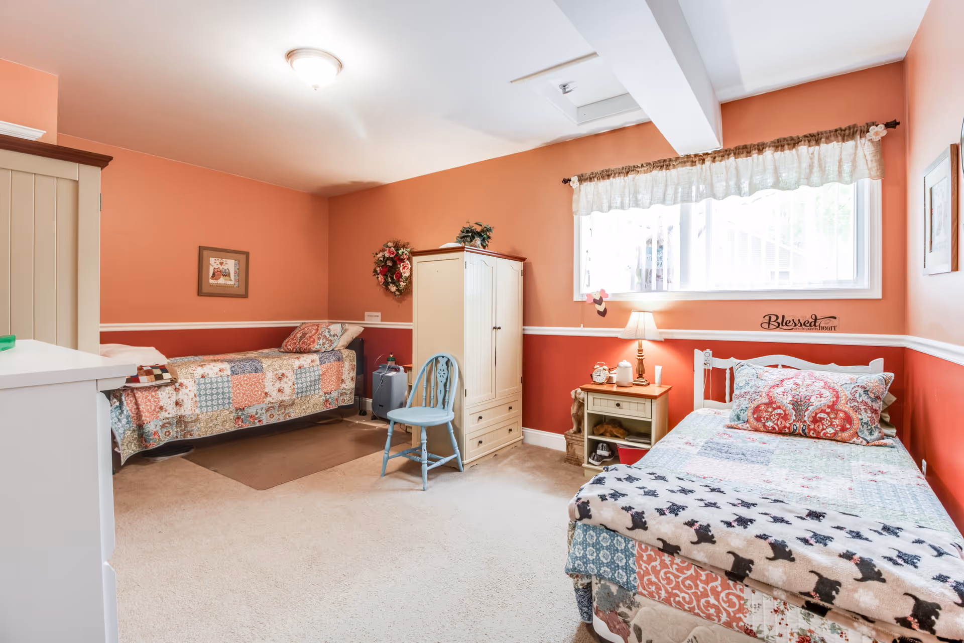 A cozy coral-painted bedroom with two twin beds dressed in quilted bedspreads, a wardrobe, nightstand with lamp, and a window.