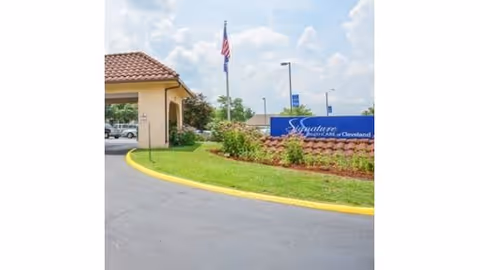 Exterior view of Signature HealthCARE of Cleveland facility showing a driveway with a yellow curb, a landscaped area with grass and bushes, an American flag on a flagpole, and a blue sign with the facility name.