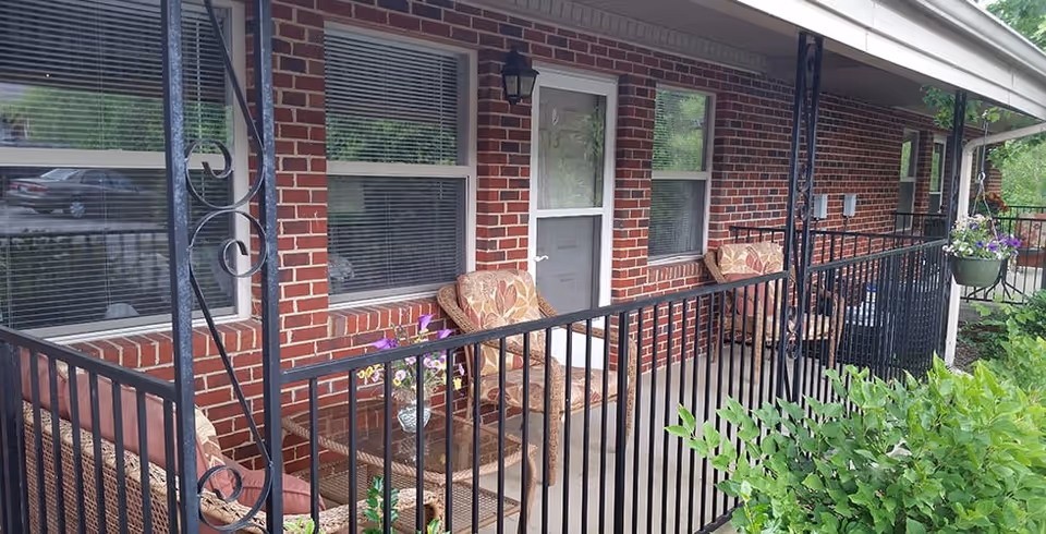 A covered outdoor patio area with brick walls, black metal railing, and several cushioned wicker chairs and a glass-top table with a flower vase. There are windows with blinds and a white door leading inside the building. Green bushes are visible in the foreground.