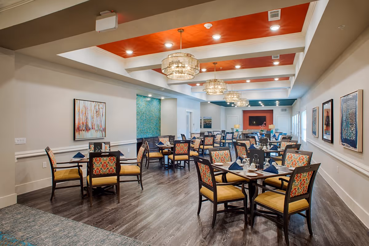 A spacious dining room in an assisted living facility with multiple tables and chairs arranged neatly. The chairs have patterned backs and yellow cushions. The ceiling features recessed lighting and decorative chandeliers with a red and white color scheme. The walls are adorned with framed artwork, and the floor is dark wood. The room has a warm and inviting atmosphere.