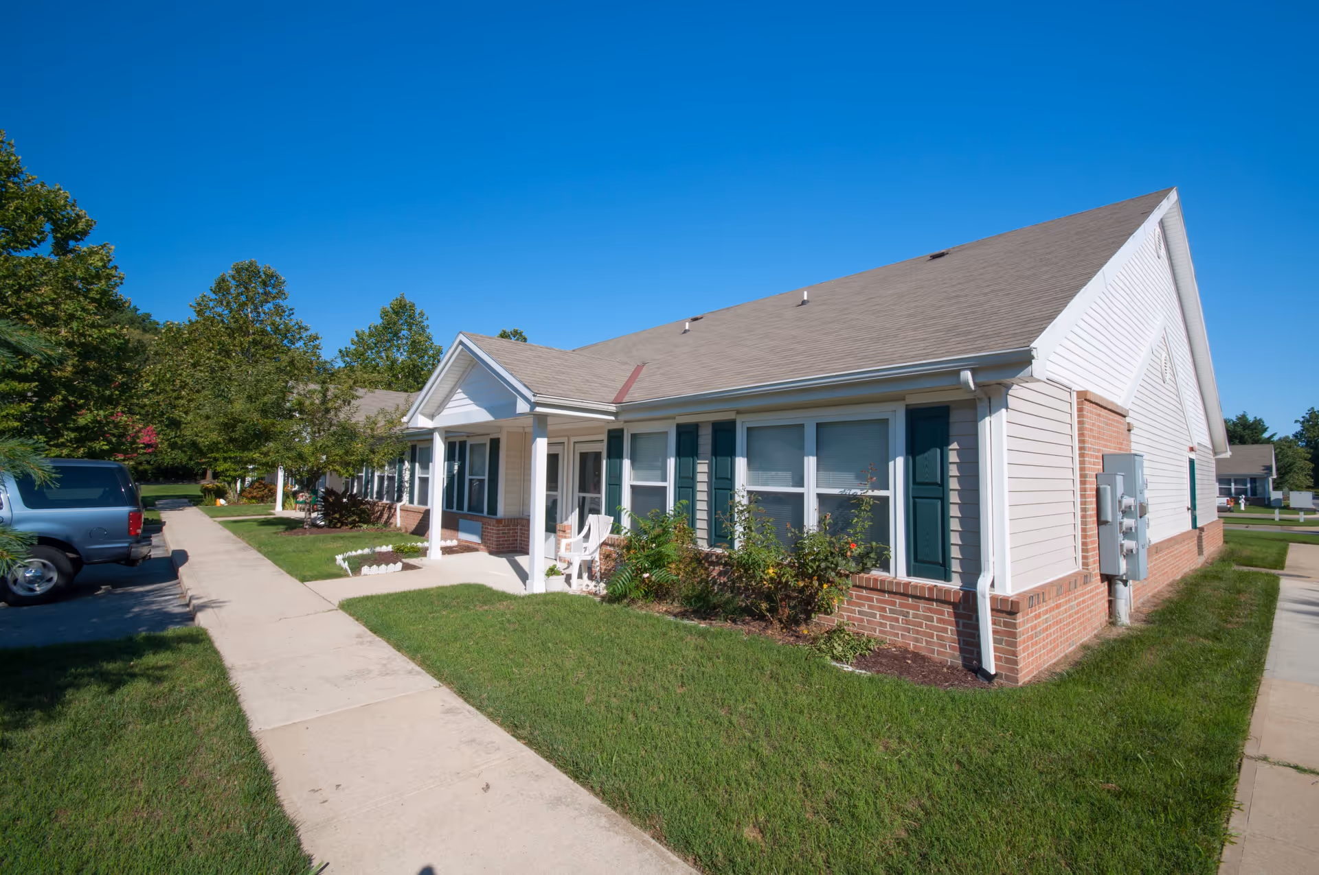 Single-story light-colored senior living building with a front porch, green shutters, lawn and sidewalk under a clear blue sky.
