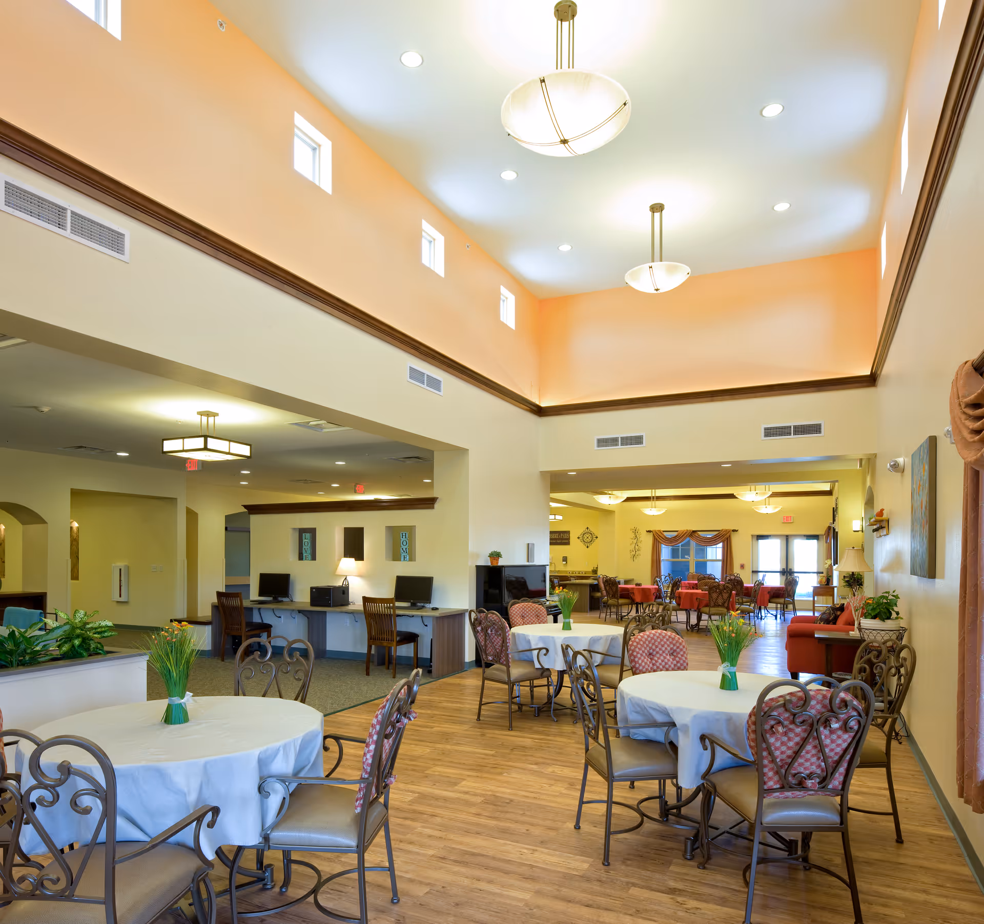 Bright senior living dining room with round tables, chairs, and warm overhead lighting.
