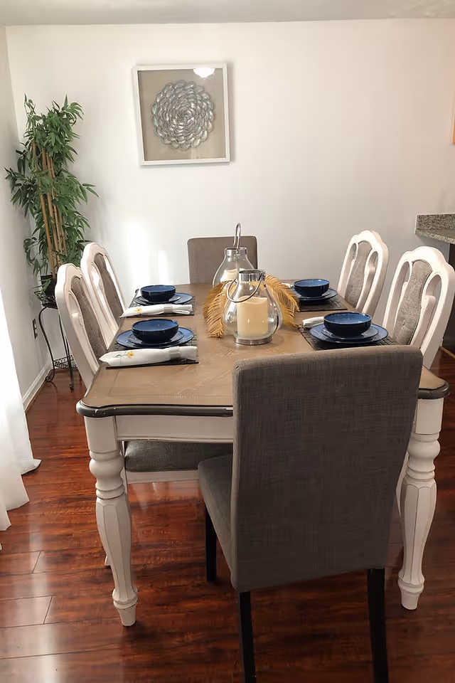 Dining room with a wooden table set for six with blue dishes, a glass candle centerpiece, and upholstered chairs on a hardwood floor.