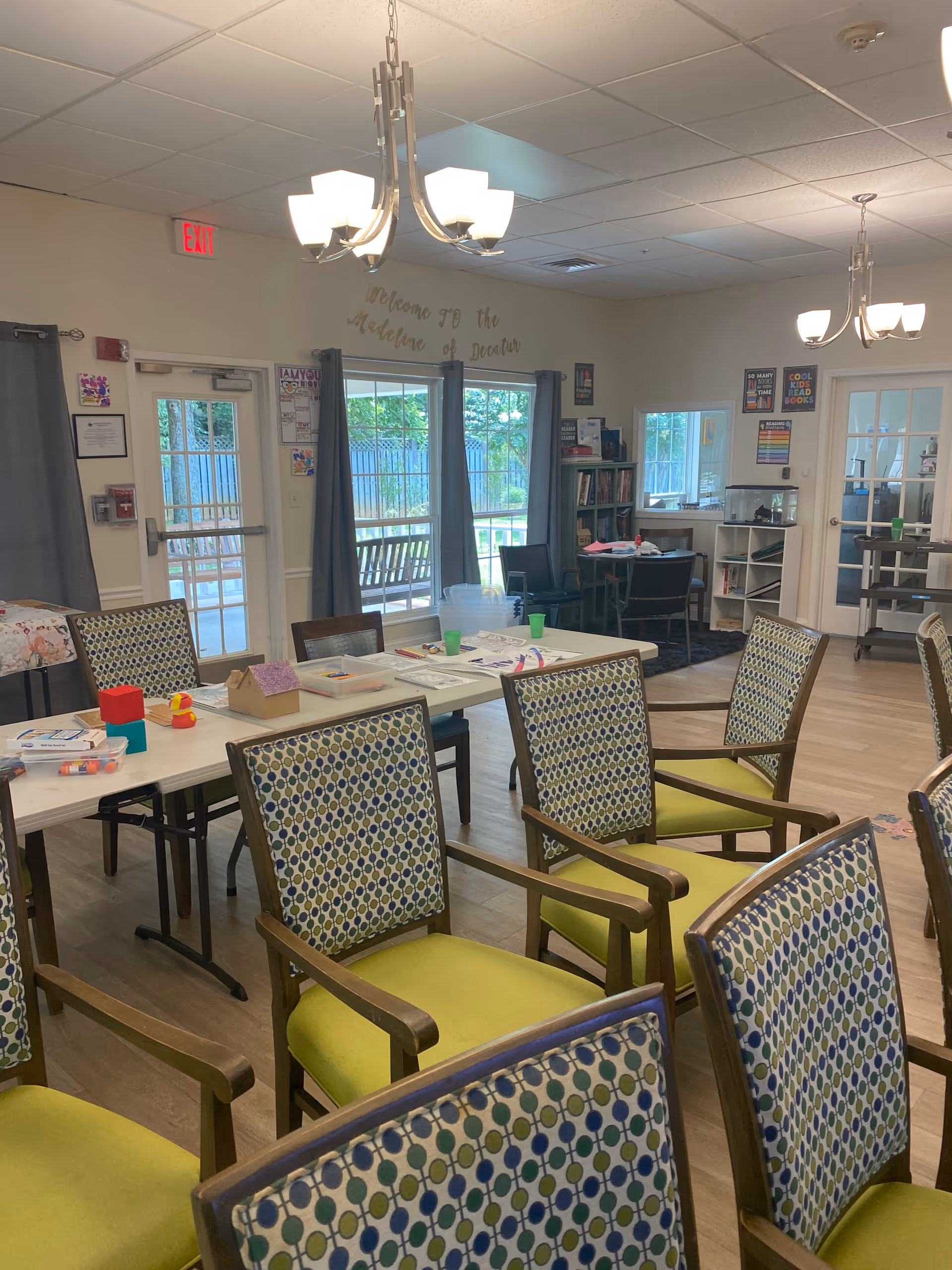 A well-lit room with multiple patterned chairs arranged around tables. The tables have various craft supplies and papers on them. There are large windows with gray curtains letting in natural light, and a door leading outside. The walls have some posters and a sign that reads 'Welcome to the Medina of Decatur.' The room has wooden flooring and modern ceiling lights.