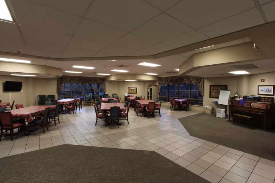 A spacious dining room in a senior living facility with multiple tables covered in red tablecloths and surrounded by chairs. The room has large windows with patterned curtains, a piano on the right side, a wall-mounted TV on the left, and framed pictures on the walls. The floor is tiled with a carpeted area near the piano.