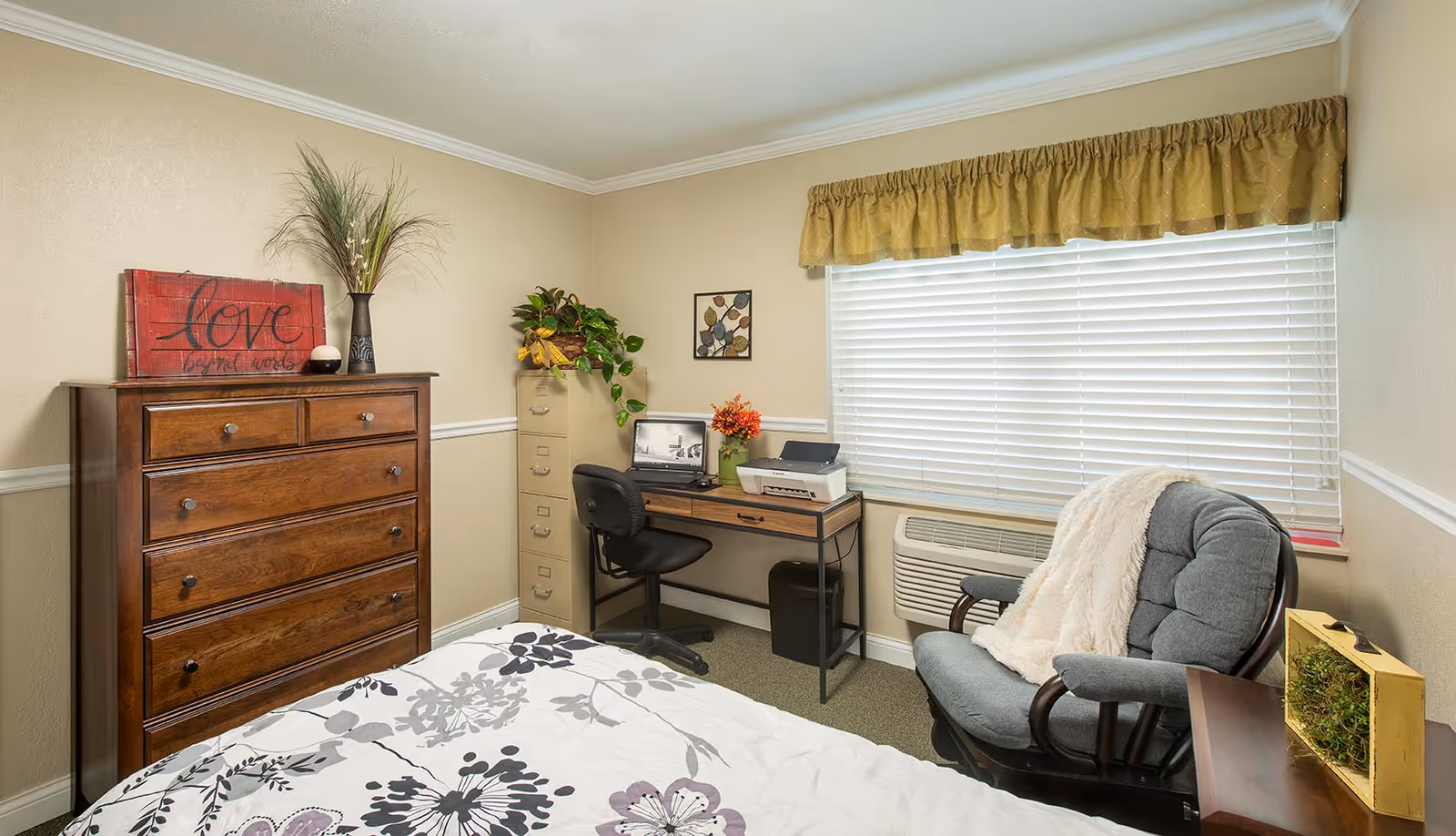 A cozy bedroom in a senior living facility featuring a bed with floral patterned bedding, a wooden dresser with a decorative sign and vase on top, a small desk with a laptop, printer, and a potted plant, a filing cabinet with a plant on it, a comfortable cushioned chair with a white throw blanket, and a window with blinds and a yellow valance.