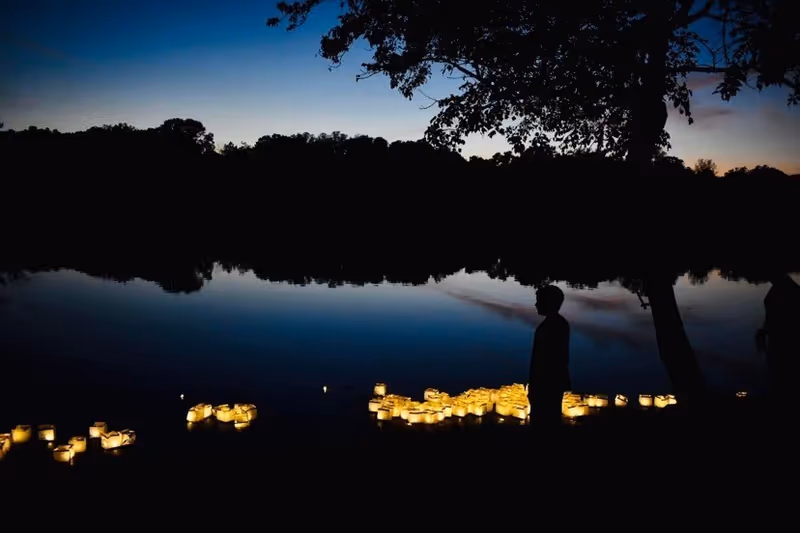 Silhouetted figures by a lake at dusk with glowing lanterns along the shore.