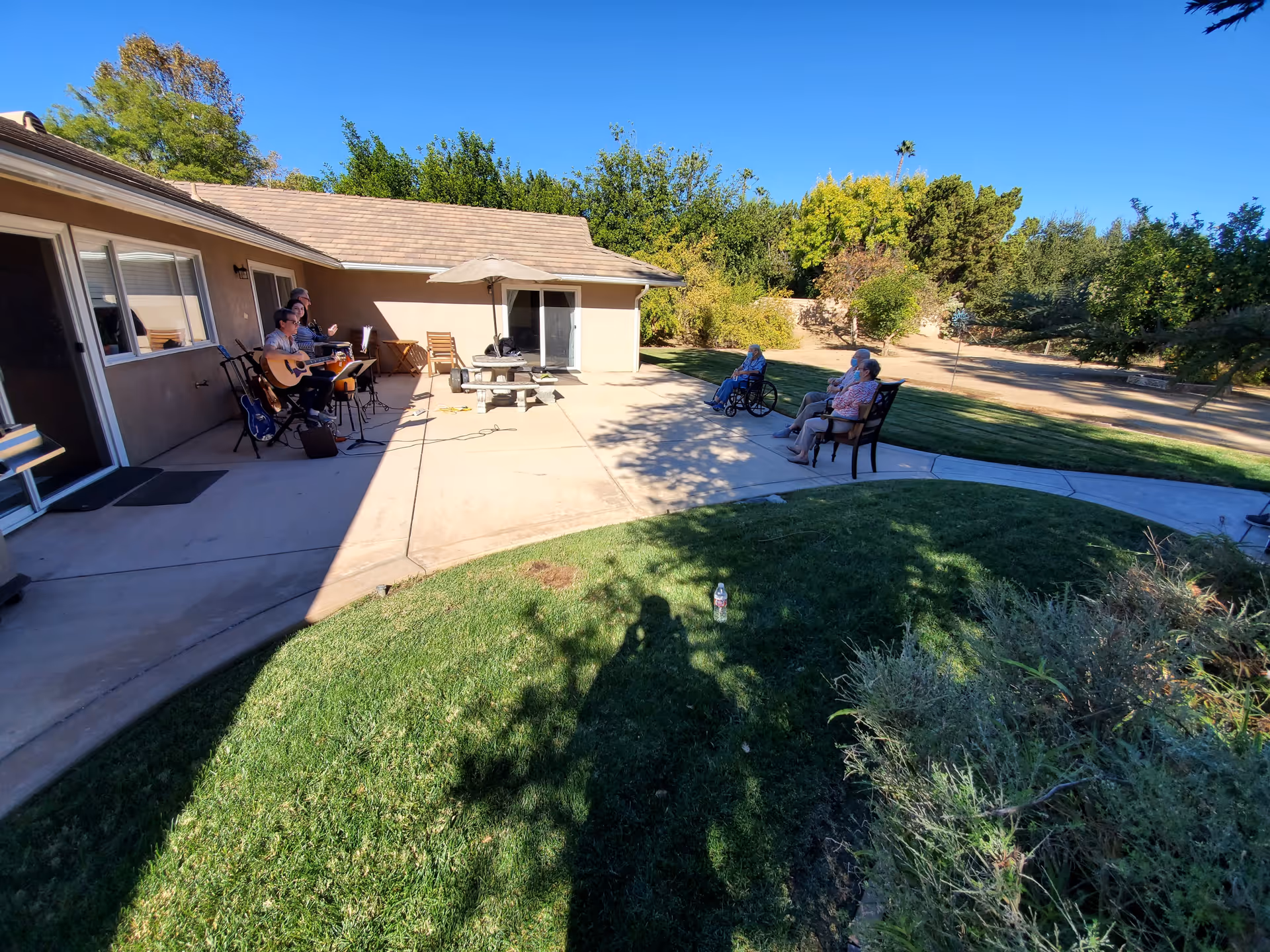 Outdoor patio area of a senior living facility with a few elderly people sitting on benches and in a wheelchair, listening to musicians playing guitars near the building. The area is surrounded by green grass, trees, and shrubs under a clear blue sky.