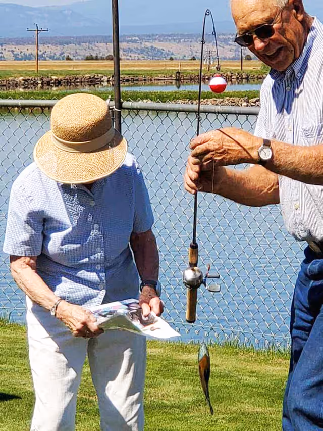 An elderly man and woman fishing by a lake. The man is holding a fishing rod with a small fish caught on the line, while the woman wearing a wide-brimmed hat looks down at a piece of paper. They are standing on grass near a chain-link fence with water and distant hills in the background.