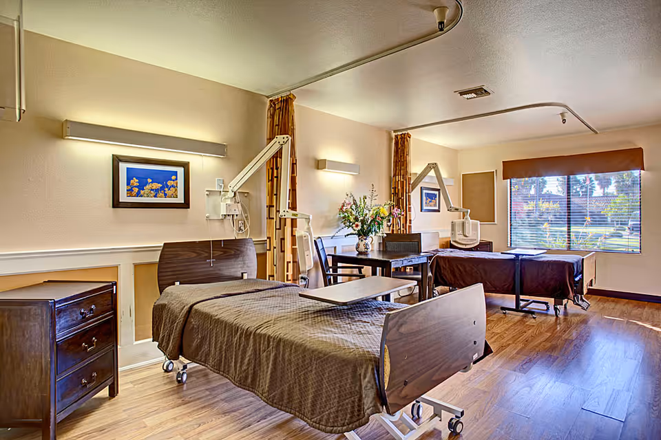 A bright and clean patient room with two hospital beds covered in brown blankets, each equipped with adjustable overbed tables and medical equipment mounted on the walls. There is a wooden nightstand with drawers, a table with chairs, floral decorations, and a large window with blinds letting in natural light.