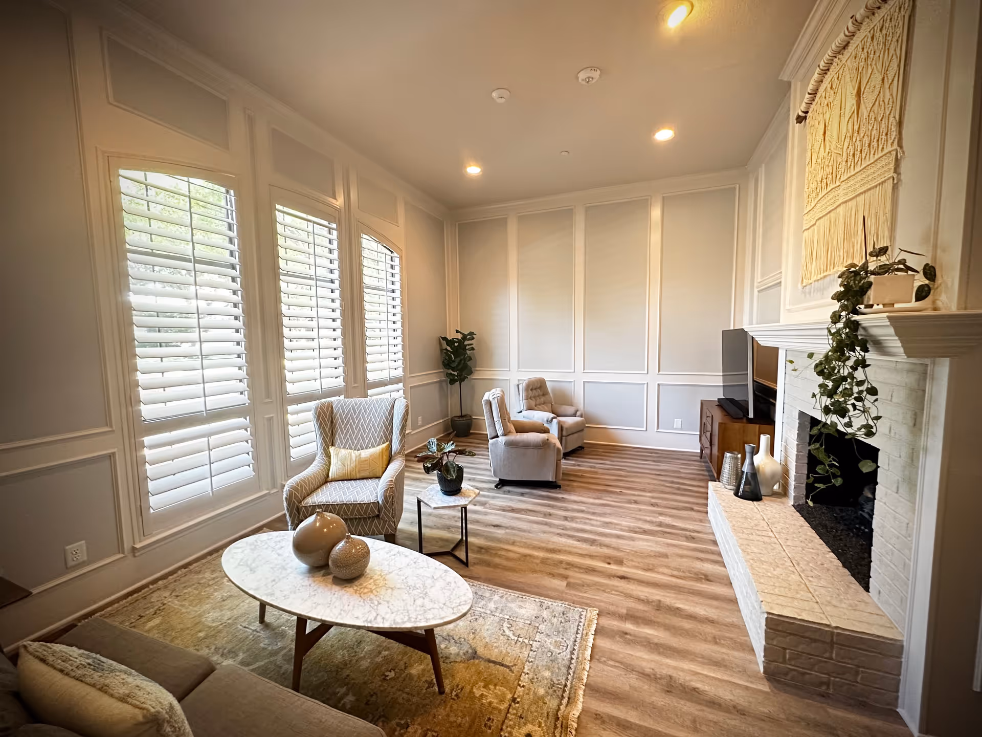 Bright living room with white paneled walls, large shuttered windows, chairs, and a fireplace.