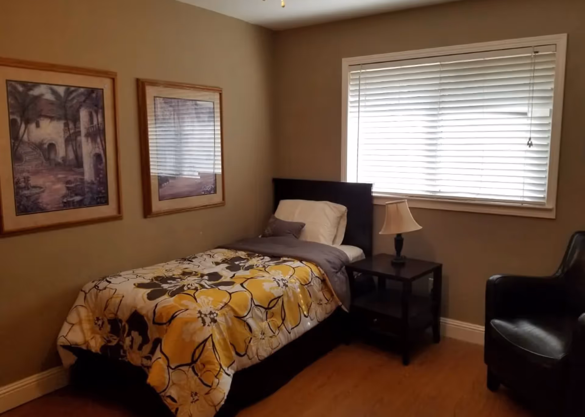 Small bedroom with a single bed dressed in yellow floral bedding, a nightstand with a lamp, framed wall art, a window with blinds, and an armchair.