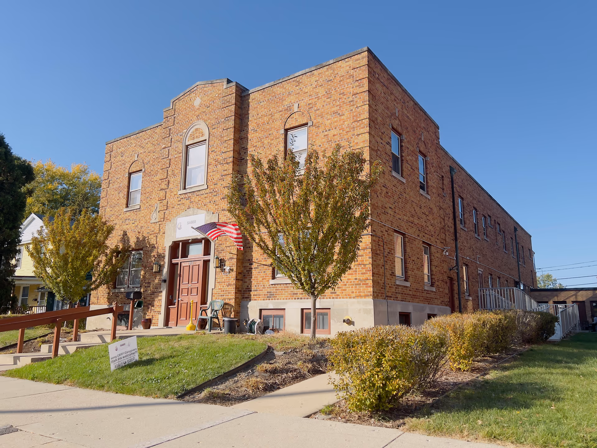 Exterior view of a two-story brick building with several windows, a main entrance with double wooden doors, an American flag mounted near the entrance, small trees and bushes in front, and a clear blue sky above.