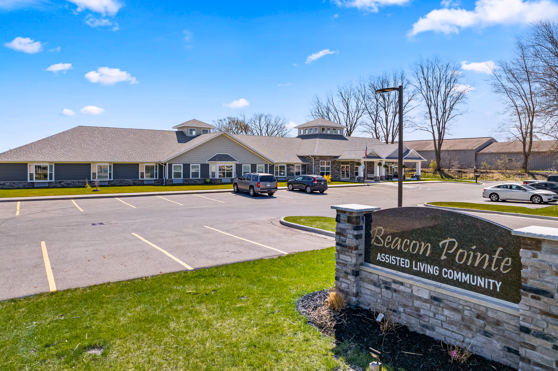 Exterior view of Beacon Pointe Assisted Living Community building with a parking lot in front, a stone sign displaying the community name, and a clear blue sky with some clouds.
