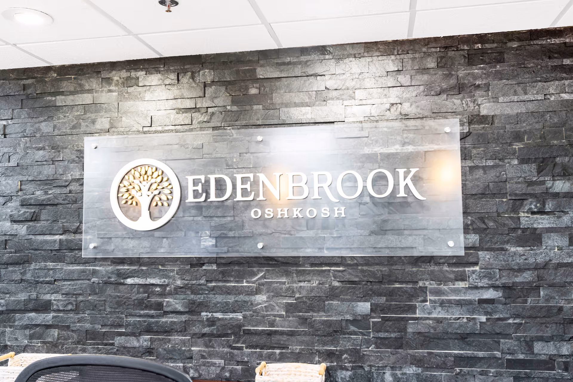 A modern interior wall with dark gray stone tiles and a clear acrylic sign mounted on it. The sign features a tree logo and the text 'EDENBROOK OSHKOSH' in white letters.