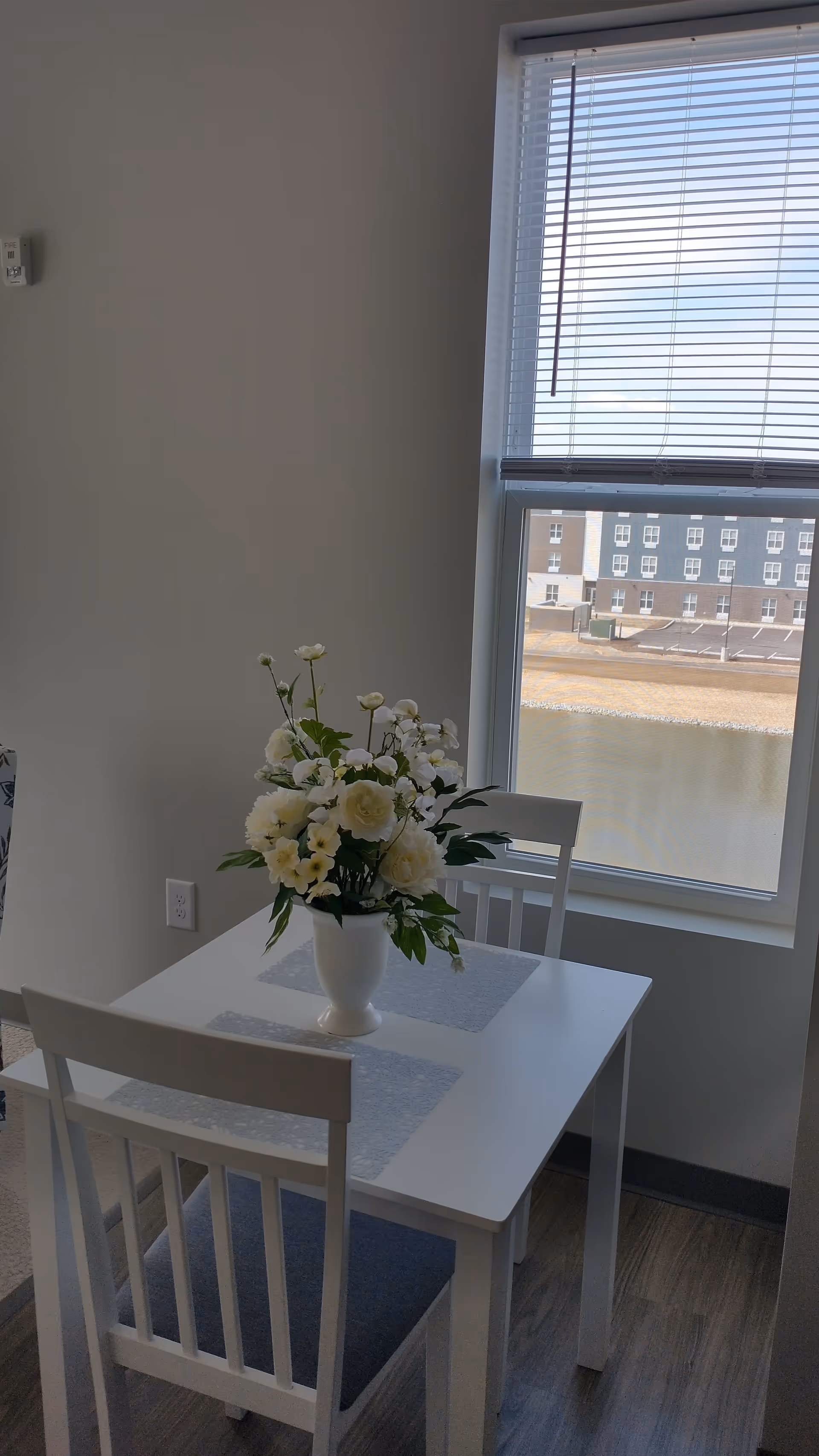 White dining table with two chairs and a vase of white flowers by a window.
