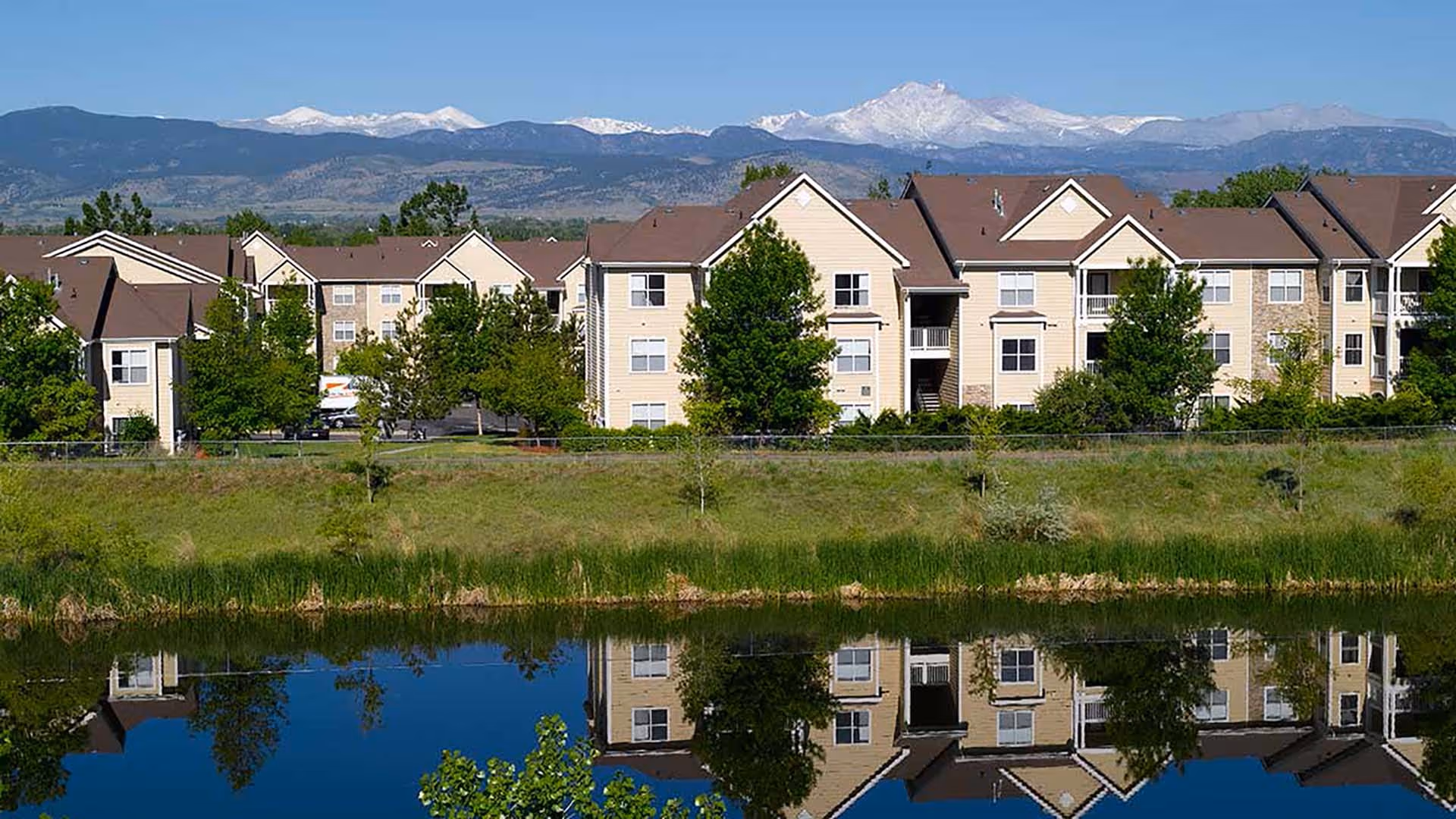 Beige multi-story apartment buildings reflected in a calm pond with a grassy shoreline and snow-capped mountains in the background.