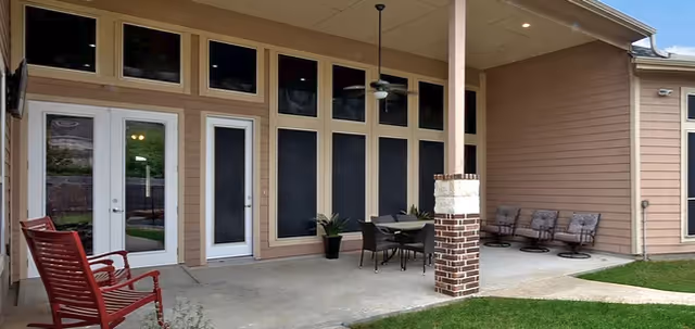 Covered outdoor patio area with a red rocking chair, a dining table with chairs, and three additional chairs lined up against the wall. The patio is attached to a building with multiple tall windows and doors, and there is a ceiling fan above the dining table. The patio opens to a grassy area.