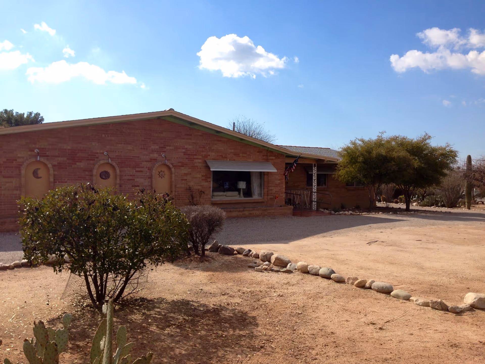 Front view of a single-story brick assisted living building with desert landscaping, shrubs, and a gravel driveway under a blue sky.