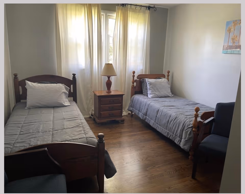 Two twin beds with a nightstand and lamp between them in a bright shared bedroom.