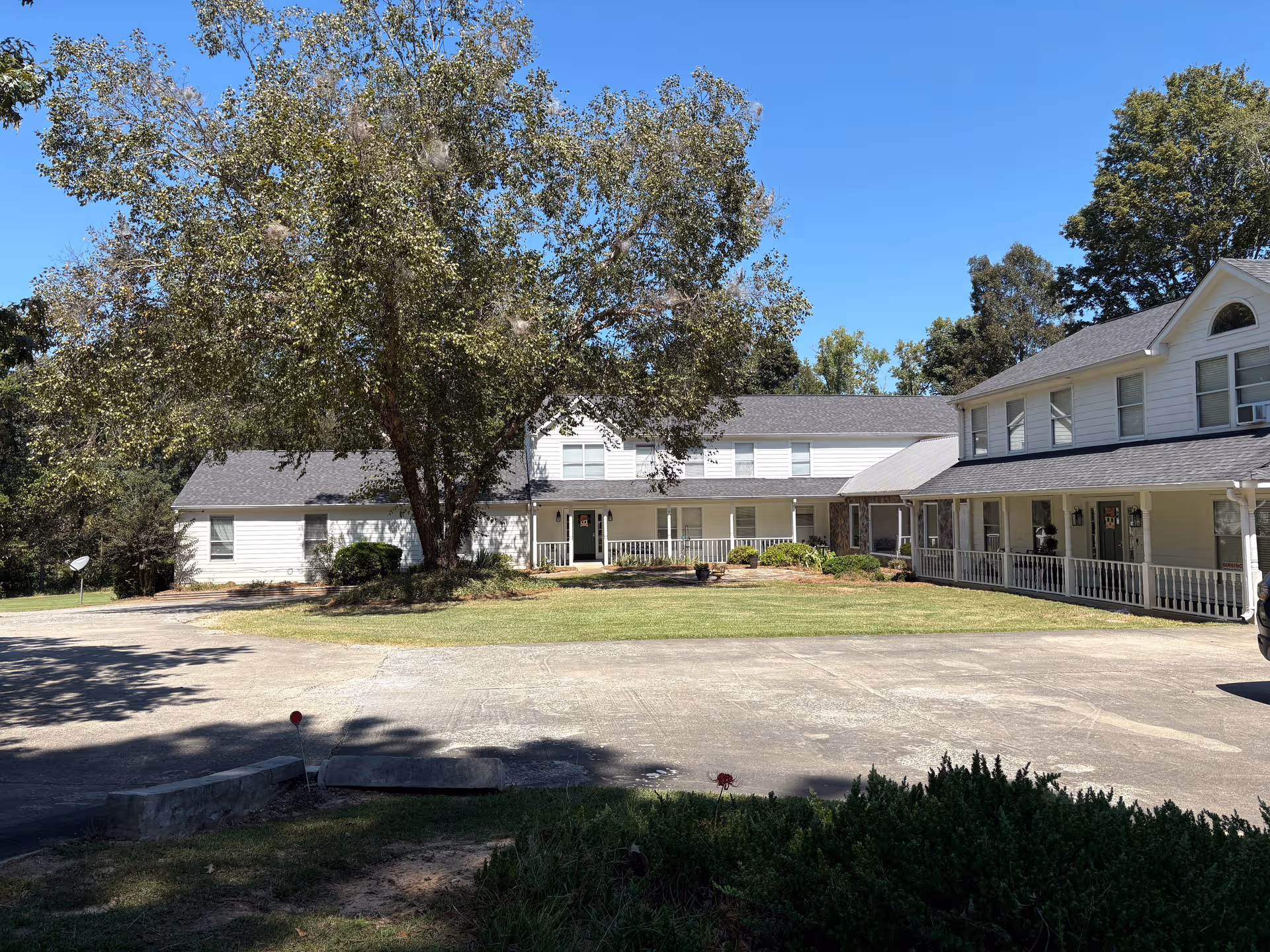 White multi-wing senior living facility with large tree, front lawn, and wraparound porch.