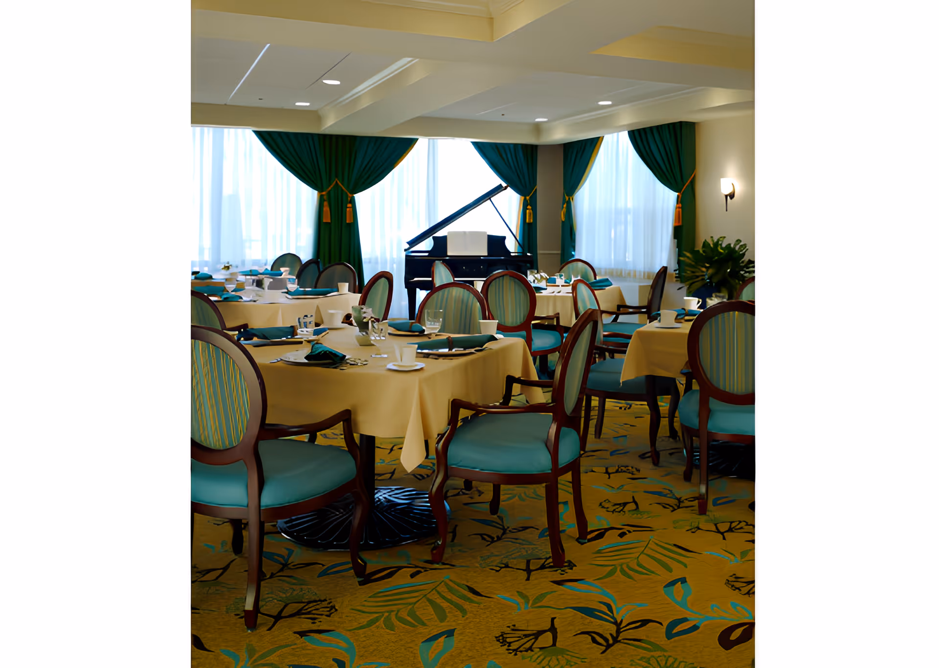 A dining room with round tables covered in beige tablecloths, set with cups, glasses, and napkins. The chairs have wooden frames with teal cushions and striped backs. Large windows with green curtains and gold tassels let in natural light. A black grand piano is positioned near the windows, and a plant is visible in the corner.
