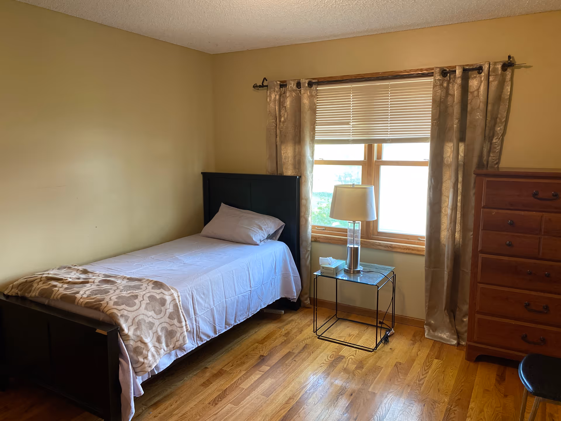 Small bedroom with single bed, nightstand, lamp, dresser, and window with curtains.