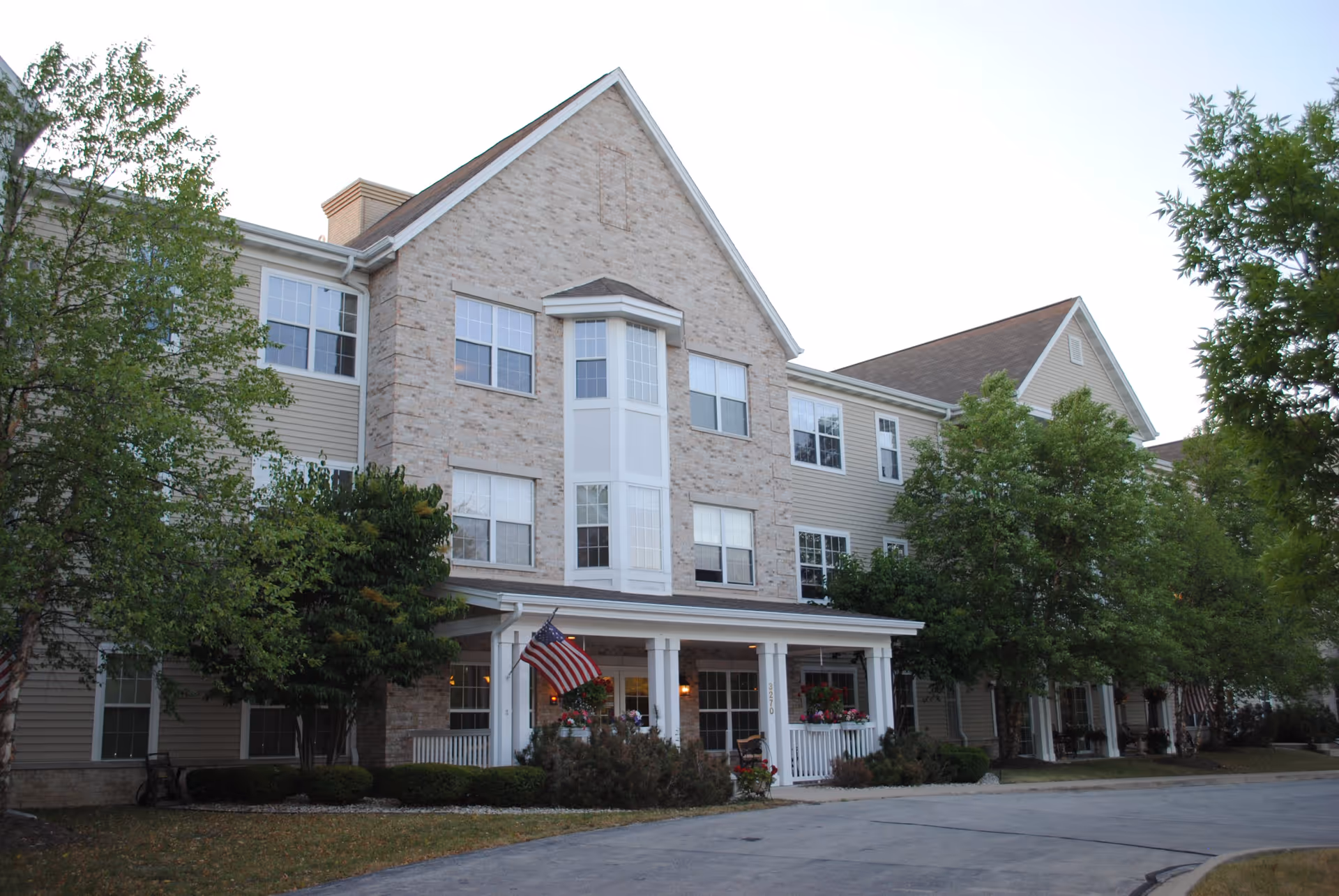 Front exterior of a three-story residential building with a covered porch entrance, an American flag, and trees along the driveway.