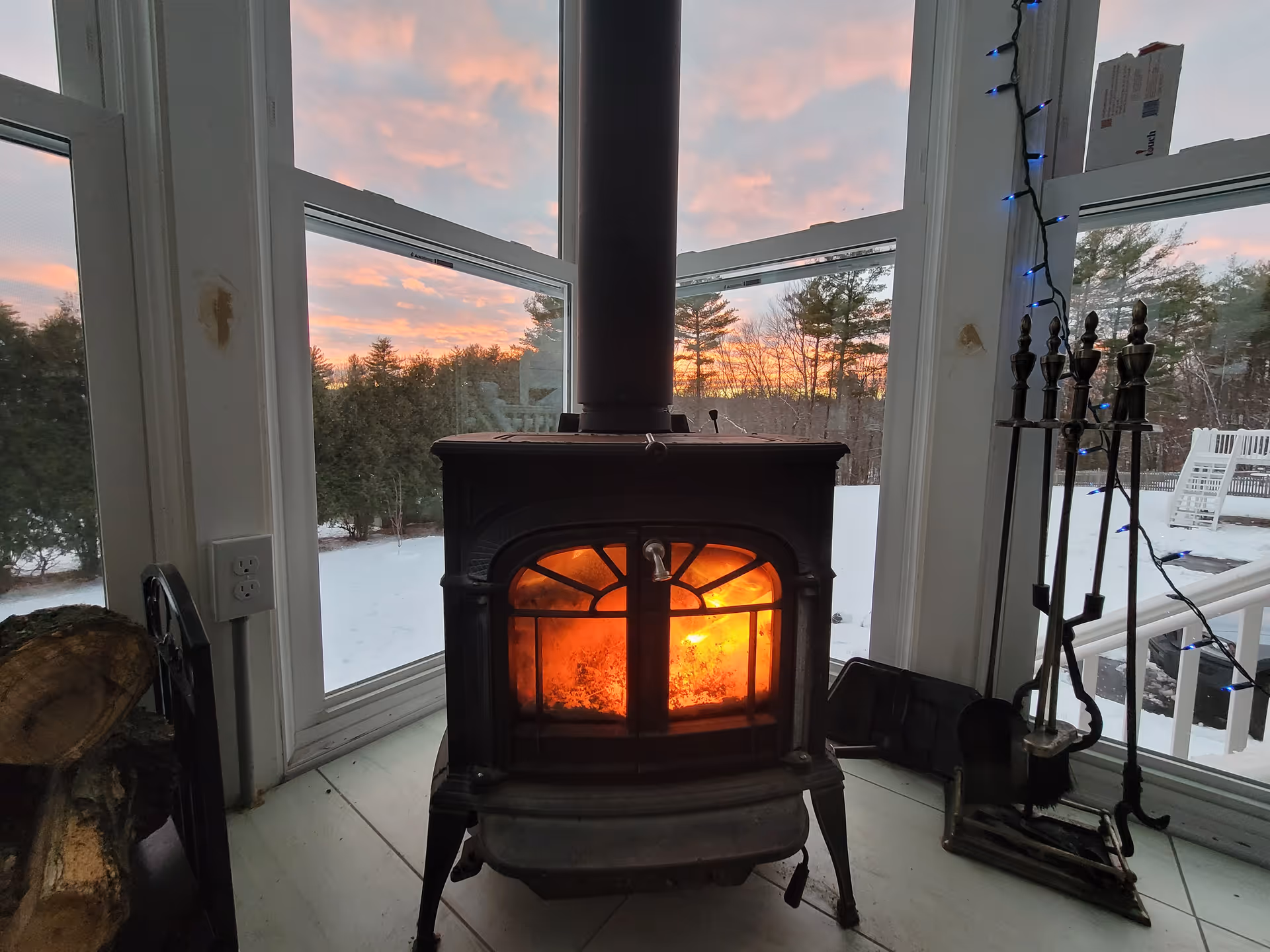 Cozy wood stove glows in sunlit interior with snowy outdoors visible through windows.