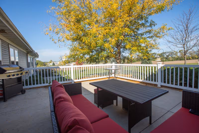 Outdoor patio with red-cushioned seating, a table, grill, white railing, and trees.