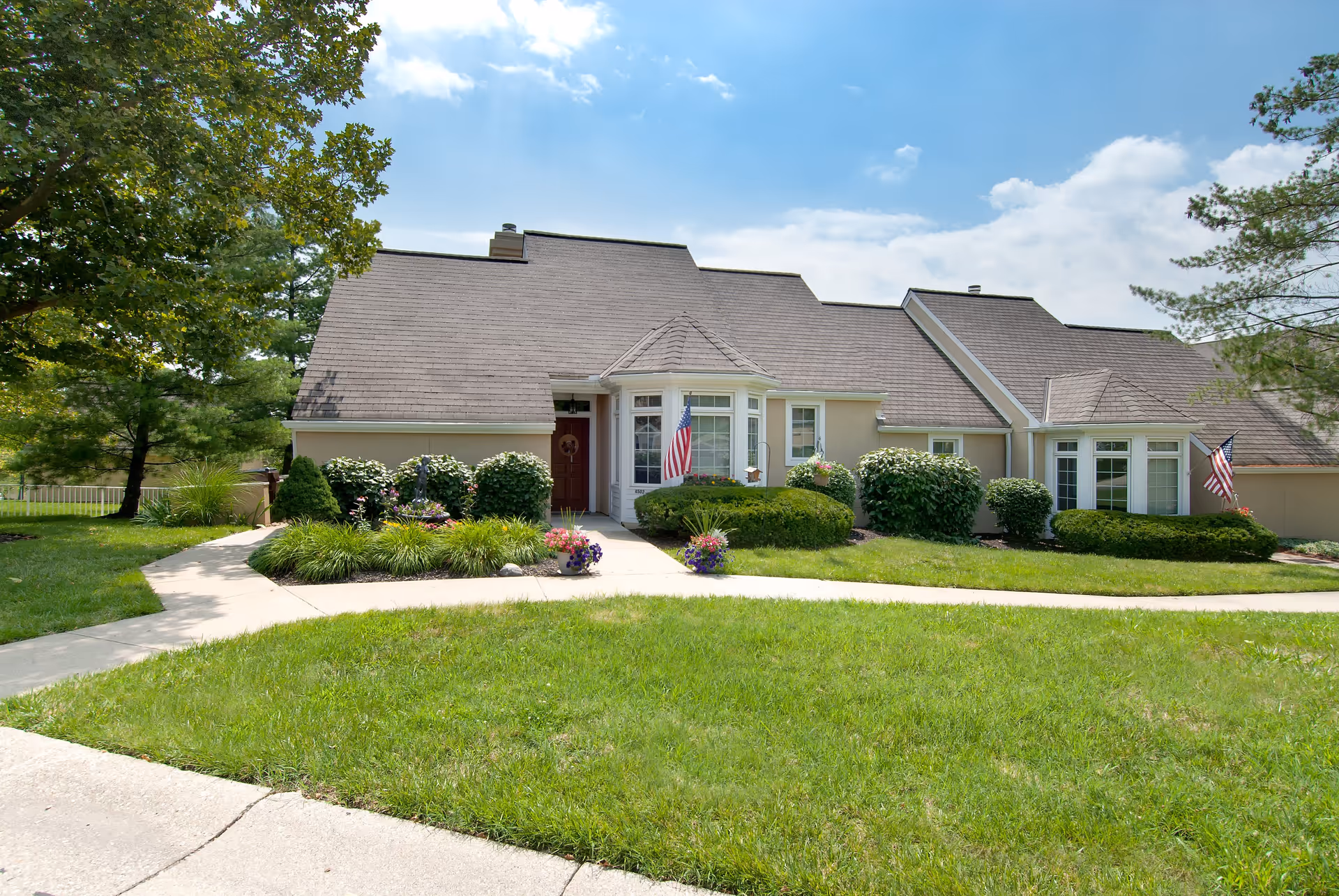 Exterior view of a single-story residential building with a sloped roof, well-maintained lawn, shrubs, and flower pots near the entrance. Two American flags are displayed near the front windows. The sky is partly cloudy with sunlight illuminating the scene.